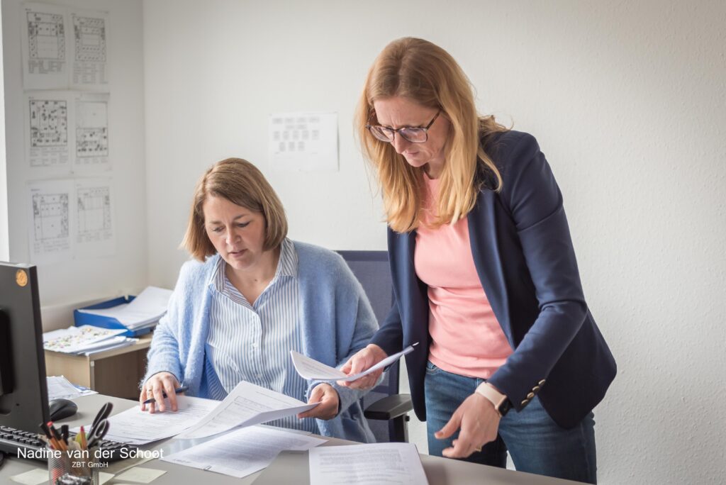 eine sitzende und eine stehende Frau an einem Schreibtisch mit Unterlagen