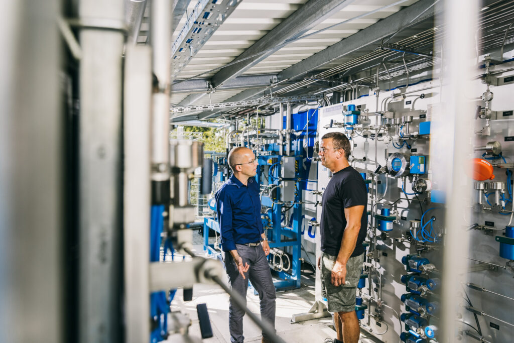zwei Personen besprechen sich vor der Hauptschalttafel der Wasserstoff-Testtankstelle auf dem Wasserstofftestfeld des ZBT