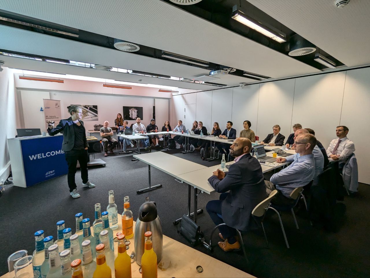 Blick in Seminarraum, Teilnehmende sitzen an Tischen, Referentin steht und gestikuliert, im Vordergrund ein Tisch mit Getränken